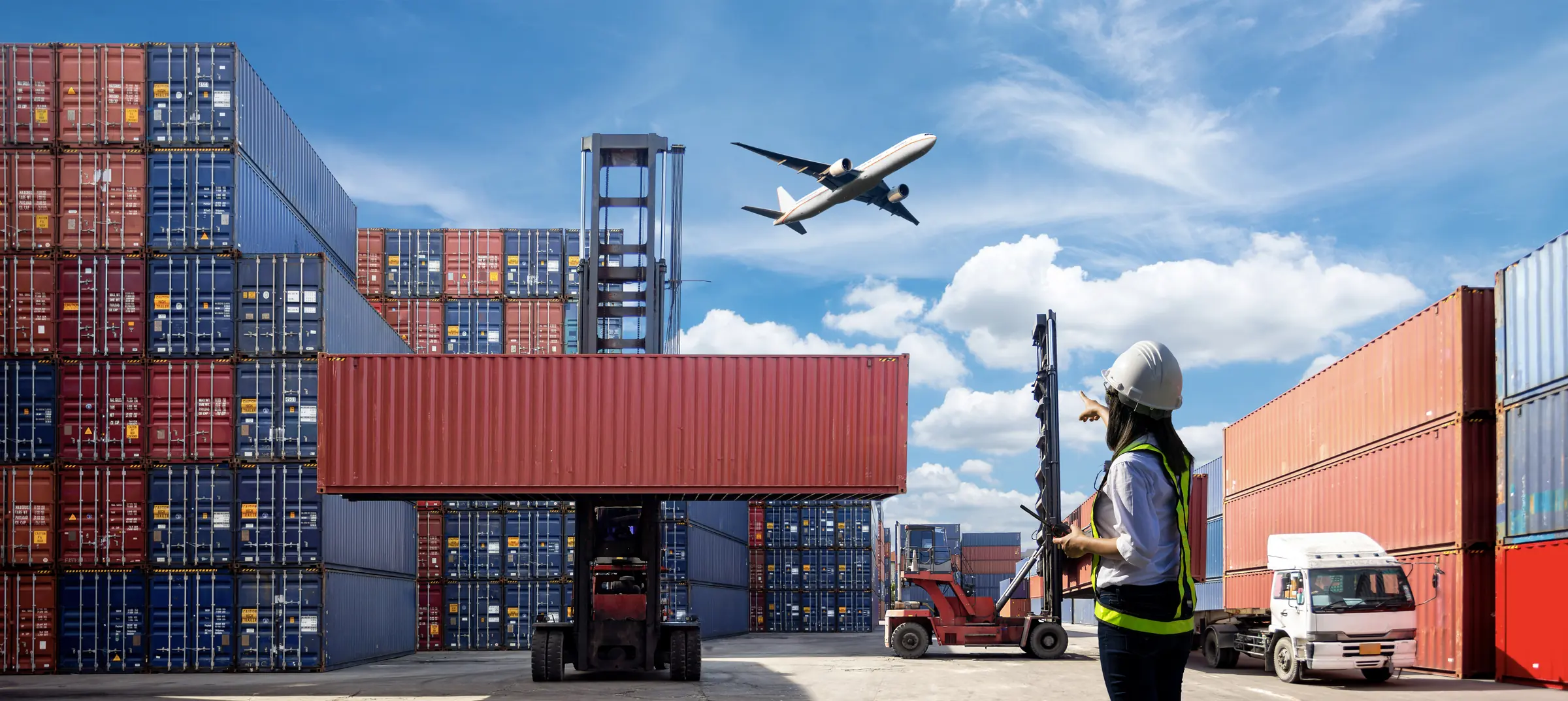 Container yard with a cargo plane overhead and a logistics worker checking shipments